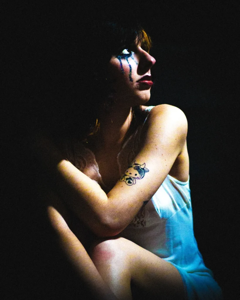 Portrait mélancolique d’une femme tatouée en chemise de nuit, assise dans l’ombre avec des larmes maquillées sous les yeux – photographie par Lo_garithme.
